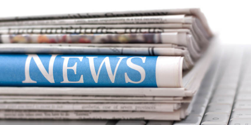 Ein Zeitungsstaper, welcher auf einer weißen Tastatur liegt. Mittig von dem Stapel ist eine Große Überschrift zu erkennen, auf der Steht News