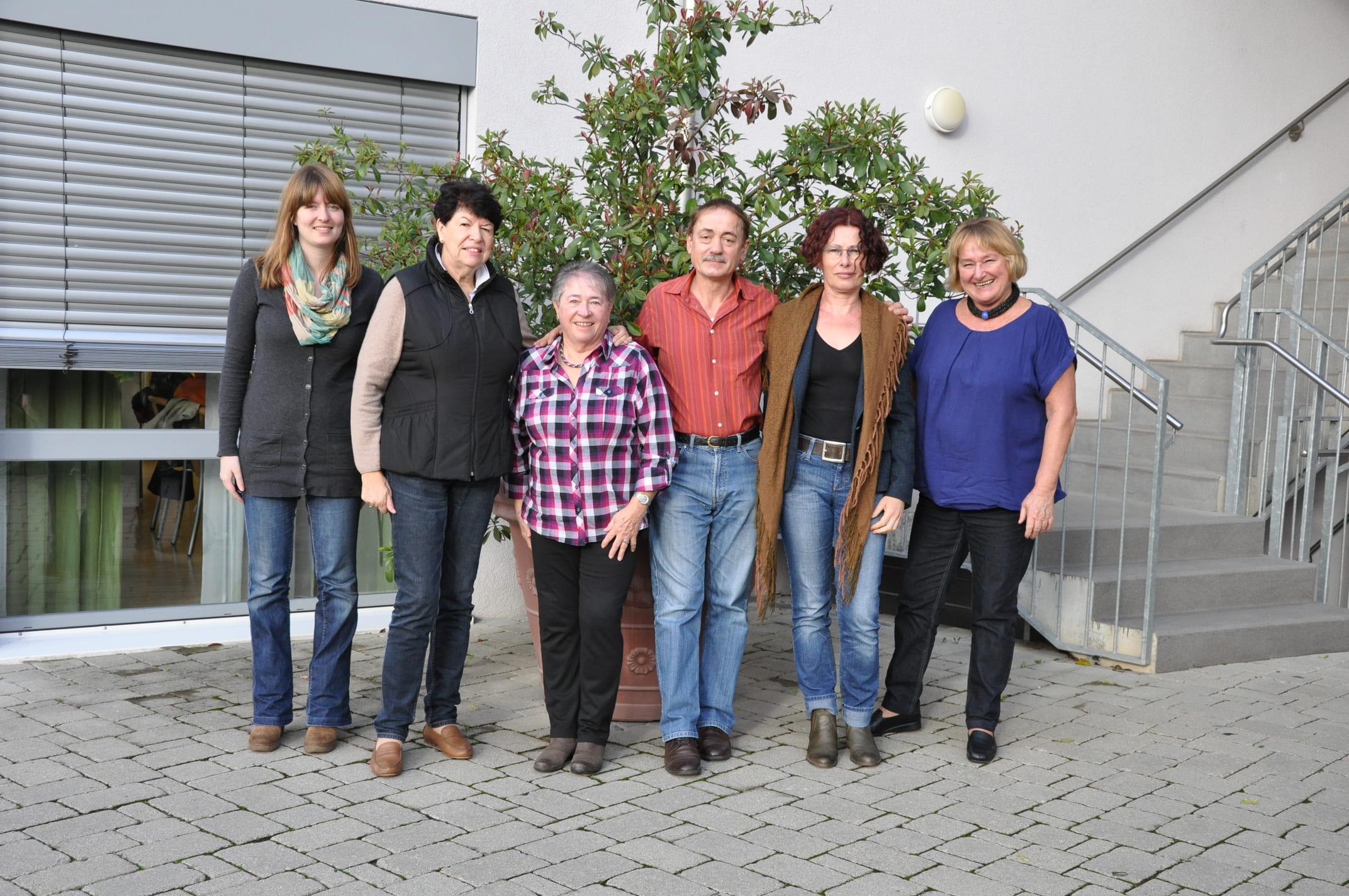 sechs Personen, die vor einem Gebäude im Freien stehen. Sie posieren nebeneinander auf einem gepflasterten Platz, hinter ihnen befindet sich eine große Pflanze in einem Topf sowie eine Treppe und Fenster mit Rollläden. Die Personen sind lässig gekleidet und lächeln