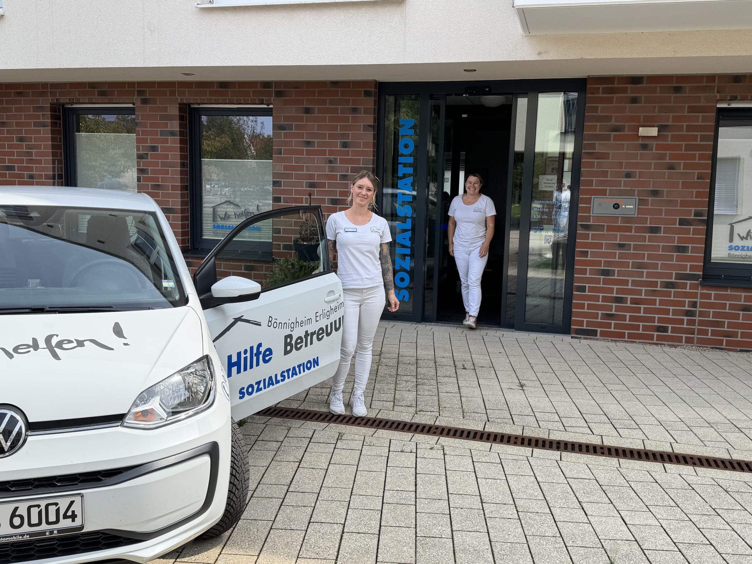 Zwei Frauen in weißer Arbeitskleidung stehen vor einem Gebäude mit der Aufschrift "SOZIALSTATION" neben einem weißen Auto mit der Aufschrift "Hilfe Betreuung SOZIALSTATION"