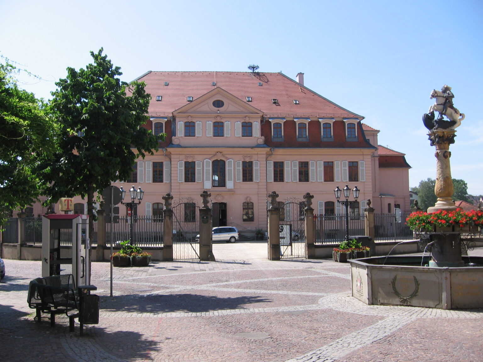 Barockgebäude mit rotem Satteldach, mehreren Fenstern und einem Tor mit Laternen im Vordergrund, rechts Brunnen mit Figur und Blumen.