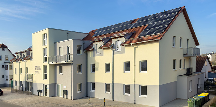 Mehrstöckiges Wohnhaus mit gelblich-beiger Fassade, mehreren Balkonen und Solarpaneelen auf dem Dach.