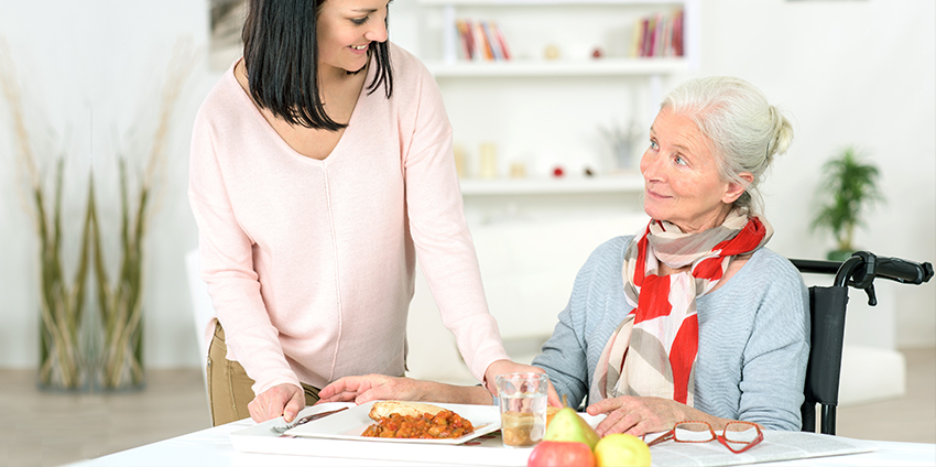 Junge Frau in rosa Pullover steht neben älterer Frau im Rollstuhl mit grauem Cardigan und rot-weißem Schal.