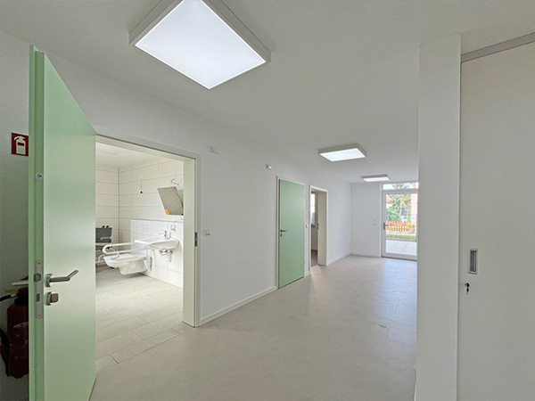 Heller Flur mit geöffneten hellgrünen Türen, links Blick in barrierefreies Badezimmer mit Haltegriffen und Duschstuhl.