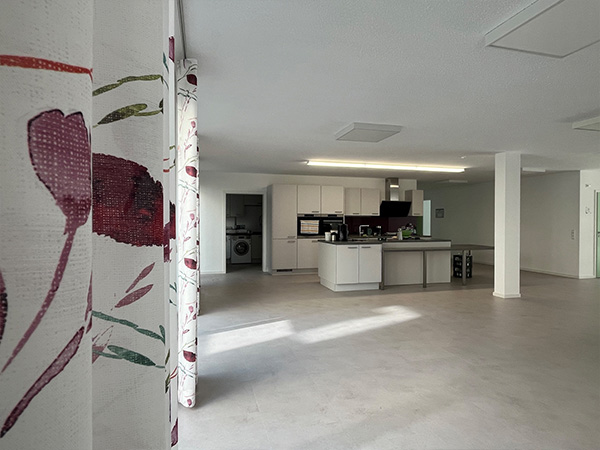 Blick in einen hellen, offenen Raum mit moderner weißer Küche und gemusterten Vorhängen im Vordergrund.