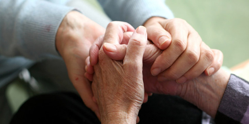Zwei Hände halten sich fest, eine ältere Hand umschließt eine jüngere Hand, beide Hände sind sichtbar.