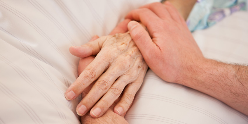 Jüngere Hand hält ältere Hand, beide auf weißer Bettdecke liegend.