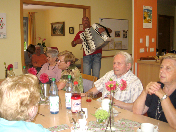 In einem hellen Aufenthaltsraum sitzen mehrere ältere Menschen an einem großen Tisch mit Getränken, während ein Mann im Hintergrund Akkordeon spielt.