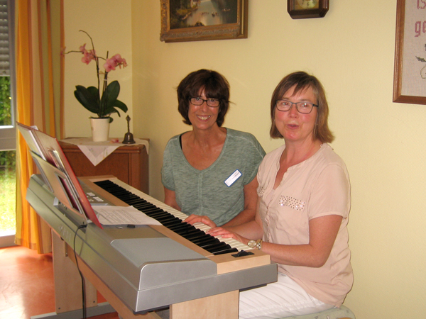 Zwei Frauen sitzen nebeneinander am Keyboard in einem gemütlichen Raum und schauen Richtung Kamera