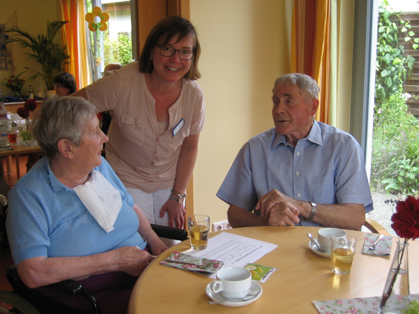 Eine Pflegerin steht bei zwei älteren Personen am Tisch, alle sitzen im hellen Wohnzimmer mit gedecktem Tisch, Getränken und Karten.