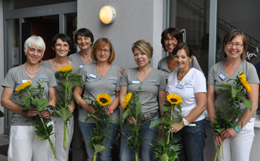 Eine Gruppe von acht Teammitgliedern, überwiegend Frauen, steht vor einem Gebäude und hält jeweils eine Sonnenblume in der Hand.