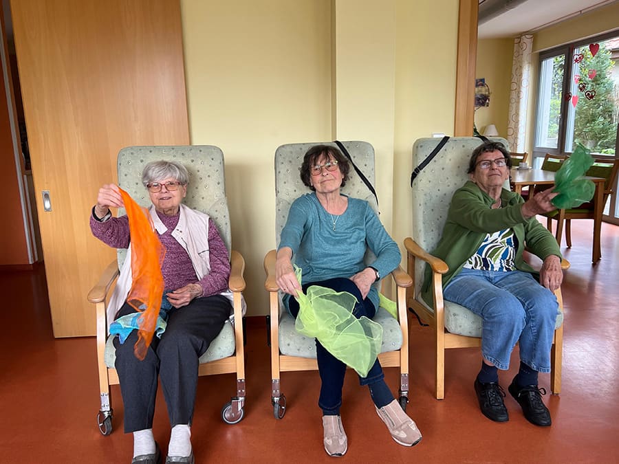 Drei ältere Frauen sitzen auf drei Stühlen und wedeln mit jeweils mit der Hand ein farbiges dünnes Tuch