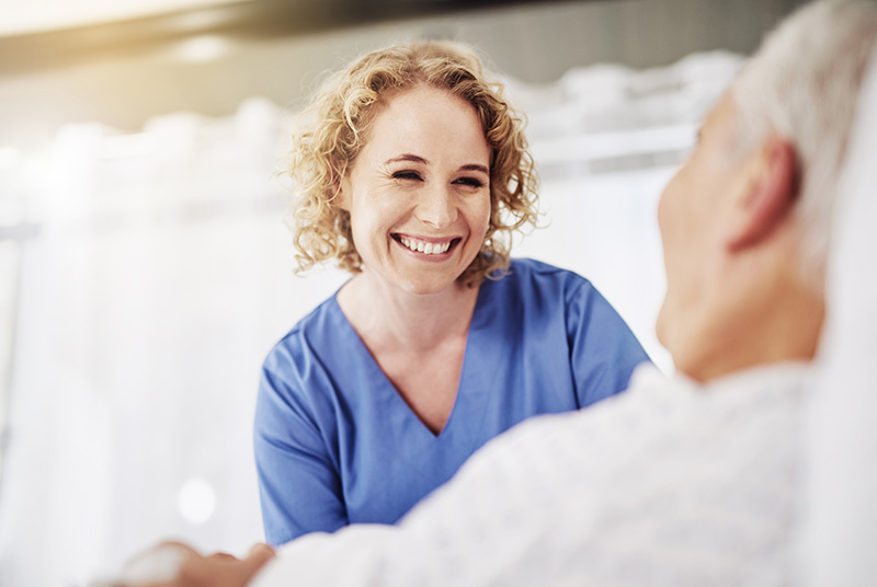 Pflegekraft mit blauem Kittel hält die Hand eines sitzenden älteren Mannes